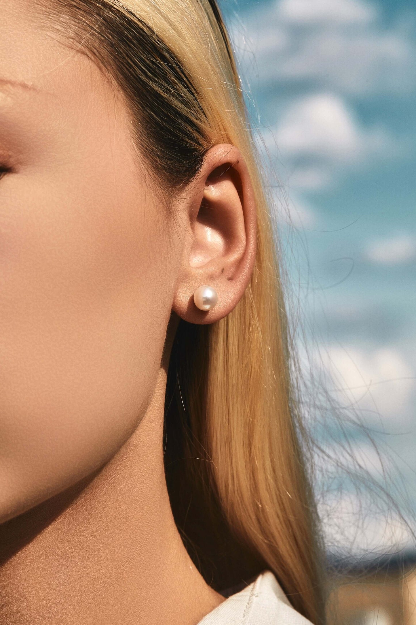 Close-up of woman wearing "Aurora" Akoya Pearl Stud Earrings by SUIHE Jewelry against a blue sky backdrop.