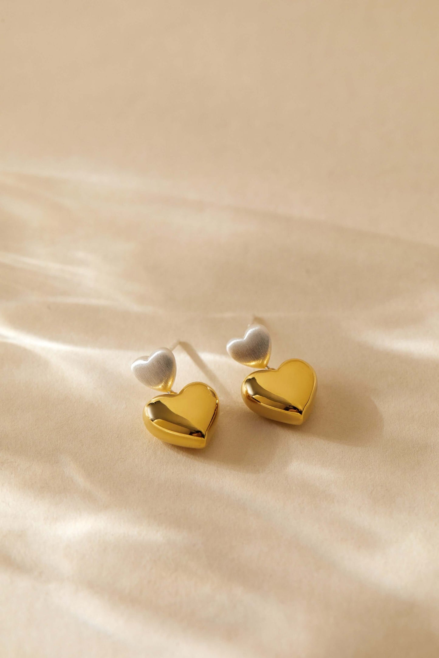 Gold and silver double heart earrings by SUIHE Jewelry on a beige background, showcasing elegant design and sophisticated craftsmanship.