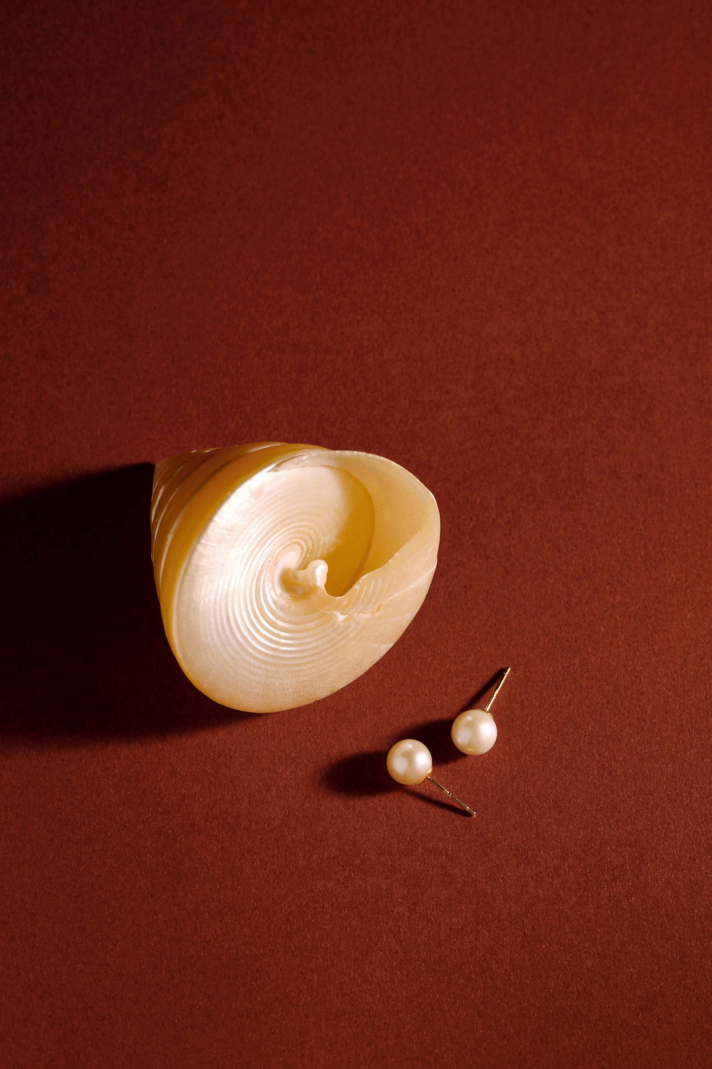 Akoya pearl stud earrings next to a shell on a muted brown background by SUIHE Jewelry.