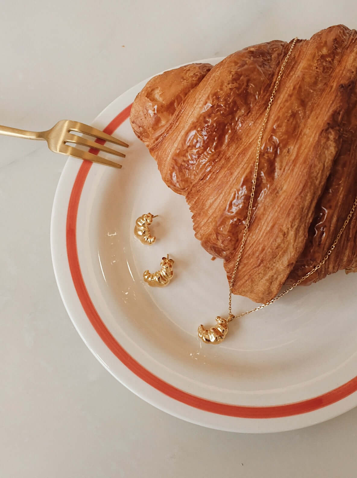 SUIHE Jewelry "Croissant" Necklace in 18k Gold Vermeil with matching earrings on a plate with a fresh croissant.