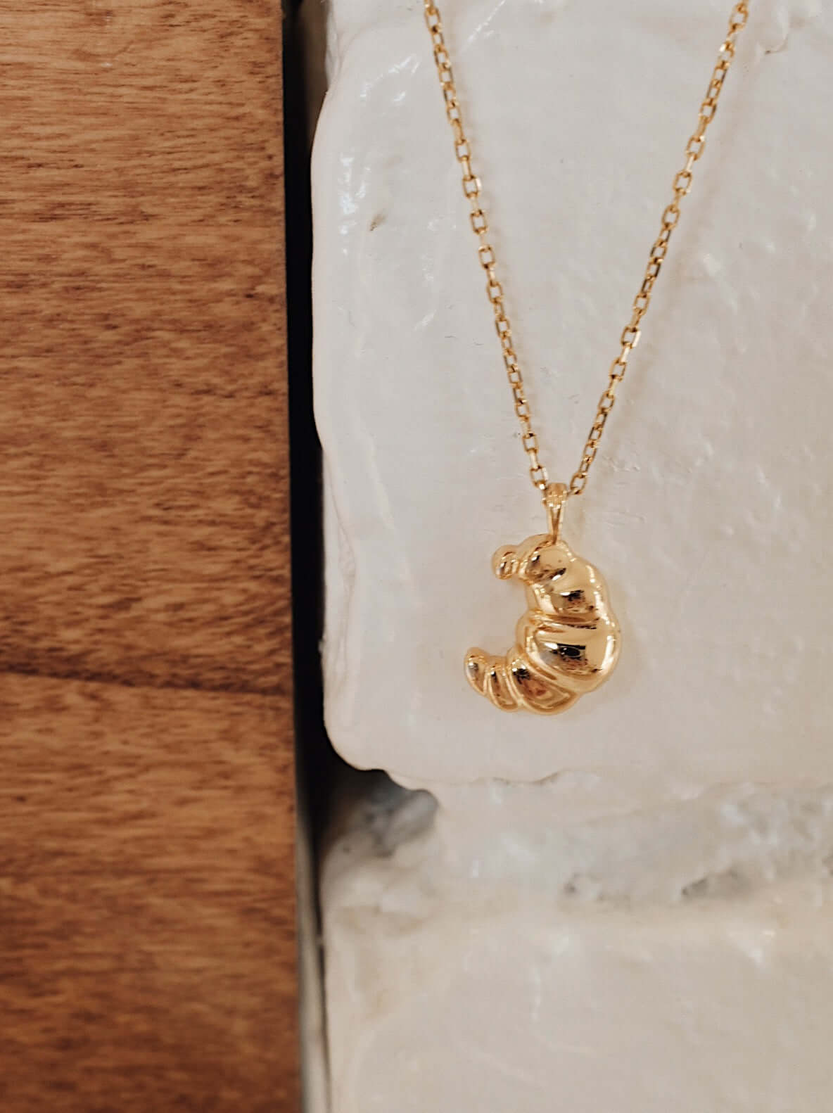 Gold vermeil croissant-shaped necklace by SUIHE Jewelry, evoking Parisian elegance with its textured design, hanging against a stone wall.