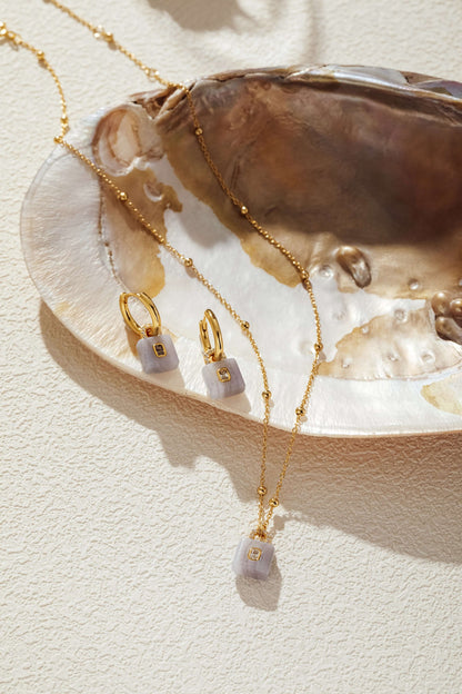 Gold-plated necklace with aquamarine lock pendant featuring a zircon, displayed with matching earrings on seashell backdrop.