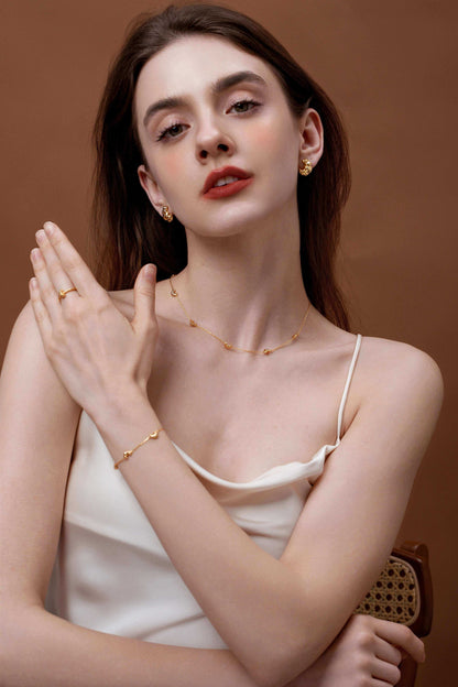 Woman wearing gold jewelry set including necklace, earrings, and bracelet with a minimalist white outfit against a brown background.