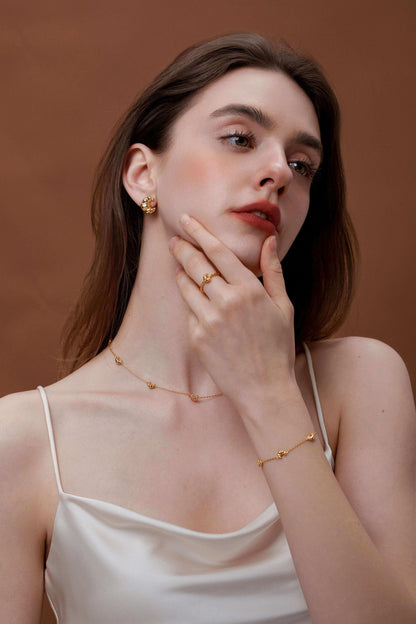 Woman wearing elegant gold jewelry including earrings, necklace, ring, and bracelet, posing gracefully against a brown background.